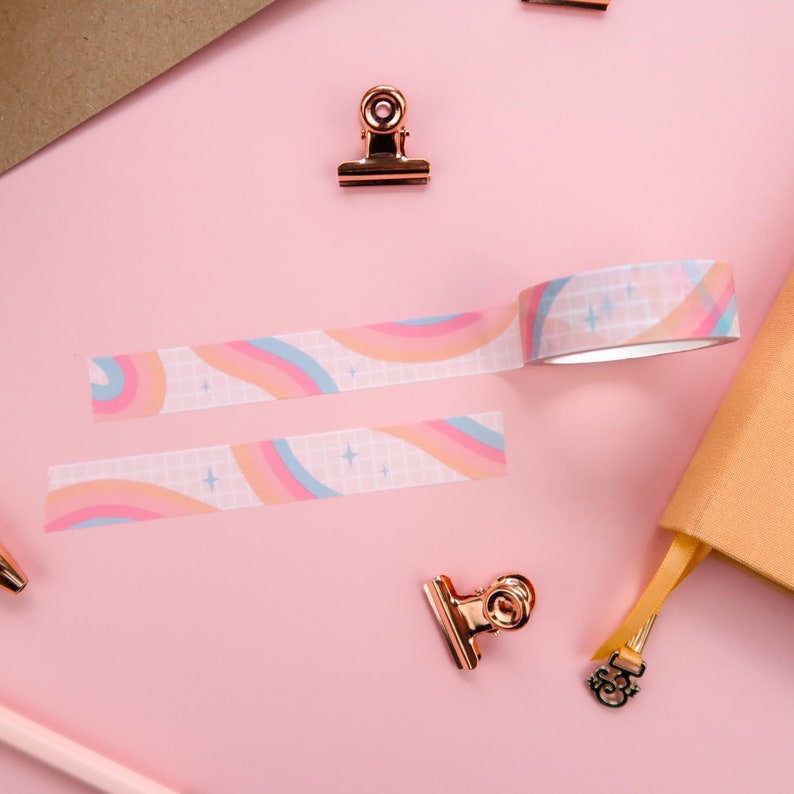 May include: Two rolls of washi tape with a rainbow and star pattern on a pink background. The tape is white with a grid pattern and features pink, blue, and orange rainbows with white stars.