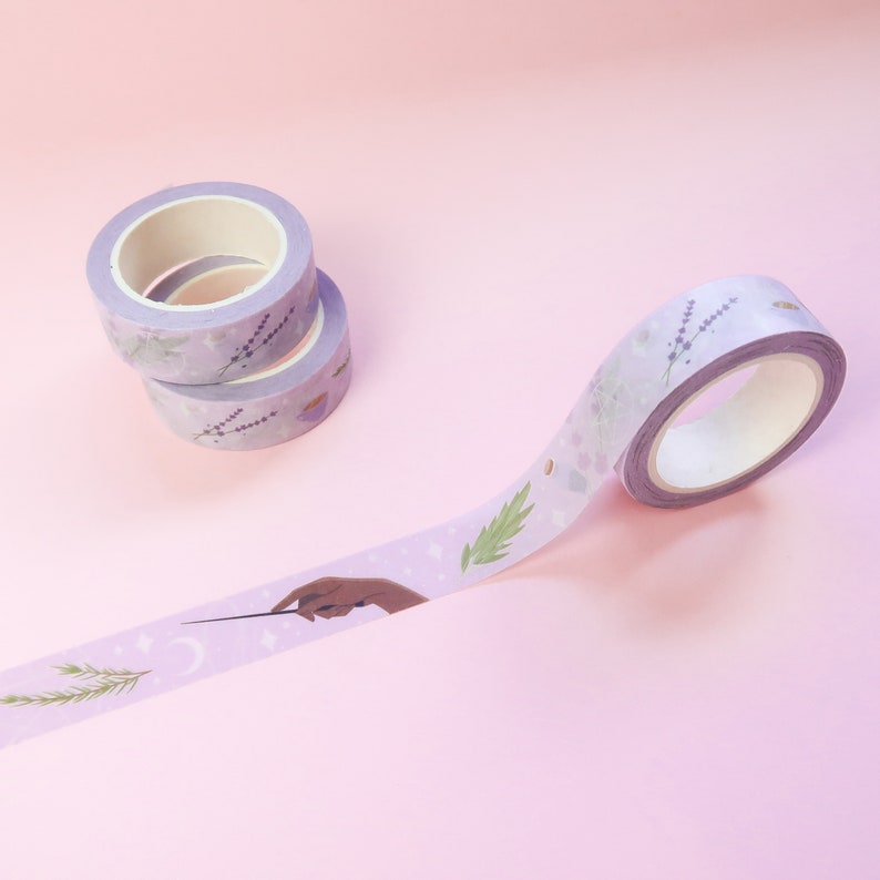 Puede incluir: Cinta washi morada con un dise&ntilde;o floral y una mano sosteniendo una varita m&aacute;gica con estrellas y una luna creciente. La cinta est&aacute; enrollada y parcialmente desenrollada sobre un fondo rosa.