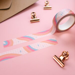 May include: A roll of rainbow patterned washi tape with a white grid background and gold stars. The tape is unrolled on a pink surface with two gold binder clips.