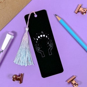 Puede incluir: Marcapáginas negro con un diseño blanco de dos manos sosteniendo un círculo de lunas y estrellas. El marcapáginas tiene un borla rosa y azul.
