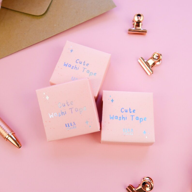 May include: Three pink boxes with the text "Cute Washi Tape" and the logo "LULA Rocks" on a pink background. The boxes are next to gold metal paper clips.