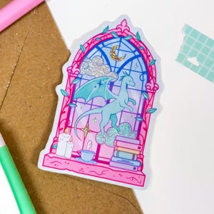 May include: A sticker featuring a light blue dragon with teal wings standing on a cloud, inside a stained-glass window design. The window has a pink border with green vines, a crescent moon, and a stack of books. Glitter accents the clouds and window.