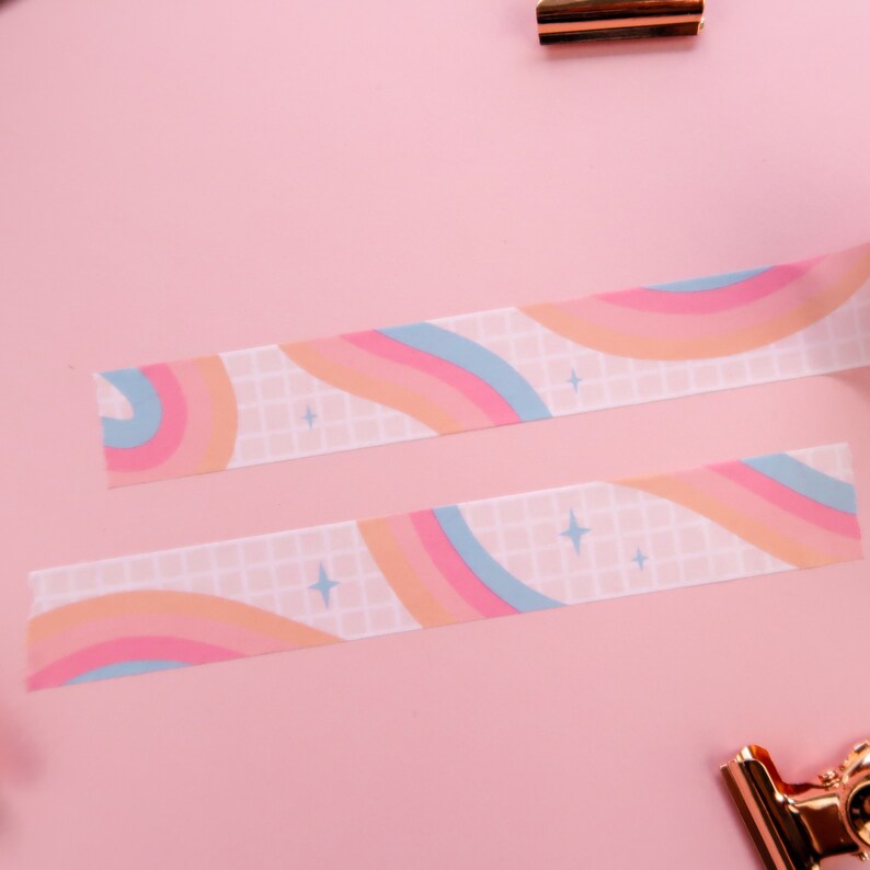 May include: Two rolls of washi tape with a rainbow and grid pattern on a pink background. The tape has a light blue, pink, and orange colour scheme with white stars.