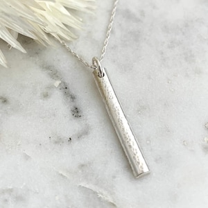 Peut inclure: Un collier avec pendentif barre en argent. Le pendentif est une barre rectangulaire texturée suspendue à une chaîne délicate. Le collier est présenté sur une surface en marbre blanc, avec une fleur blanche en arrière-plan.