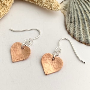 May include: A pair of heart-shaped earrings. Each earring has a hammered copper heart pendant hanging from a silver-coloured ear wire. The earrings are shown on a white surface, with a starfish and seashell in the background.