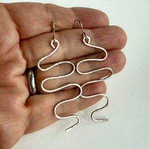 May include: Silver-coloured, abstract dangle earrings. The earrings have a unique, wavy design with a hammered texture. The hooks are simple and curved. The earrings are held in a hand against a white background.