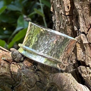 May include: A wide silver ring featuring a hammered, textured surface and a smooth, polished horizontal band running across the center. The metal has a reflective, metallic finish that catches the light, creating highlights and shadows across the band.
