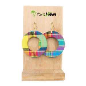 Peut inclure: Une paire de grandes boucles d'oreilles rondes et colorées avec un motif à carreaux. Les boucles d'oreilles sont en tissu et ont une fermeture à crochet dorée. Les boucles d'oreilles sont présentées sur un support en bois avec le texte "Yovo Newi" sur le devant.