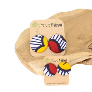 Può includere: Un paio di orecchini rotondi in tessuto colorato con un motivo bianco, rosso e giallo. Gli orecchini sono esposti su una superficie di legno con un biglietto che recita "Yoyo Newi" e "www.yoyo-mewi.com".