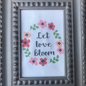May include: A framed cross-stitch with a white background and a wreath of pink and red flowers with green leaves. The text "Let love bloom" is in the center of the wreath.