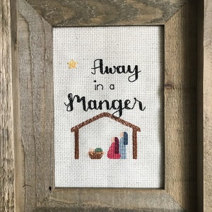 May include: A framed cross-stitch artwork with the words "Away in a Manger" in black script. The image depicts a nativity scene with a star, a manger, and figures in red and blue. The frame is made of rustic, weathered wood.