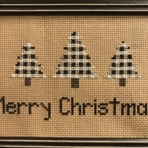 May include: Cross-stitched Christmas artwork in a black frame. Three Christmas trees with black and white checkered patterns are displayed above the words "Merry Christmas" in black. The background is a tan burlap fabric.