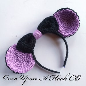 May include: A handmade crochet headband featuring large, round ears in lavender and black. The ears are attached to a black headband with a black bow and a lavender accent. The text "Once Upon A Hook CO" is at the bottom.