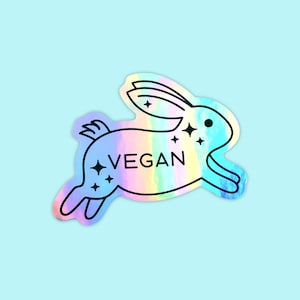 May include: A holographic sticker featuring a rainbow-colored bunny rabbit jumping with the word "VEGAN" written on its body. The bunny has stars around it.