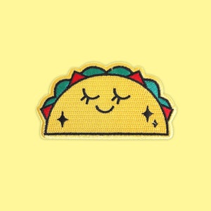 May include: A yellow embroidered taco with a smiling face and closed eyes. The taco has green and red details around the edges.