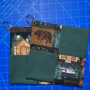 May include: Two green pot holders with a patchwork design. One pot holder features a cabin, a bear, and the words "Cabin" and "Moose". The other pot holder features a camper, trees, and the words "Lakeview".