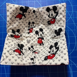 May include: A white fabric pot holder with a red border and a pattern of Mickey Mouse characters on a white background with gray Mickey Mouse ears.
