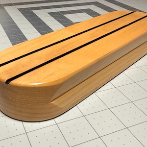 May include: An elongated, oval-shaped wooden box with a smooth, light-toned finish. The lid features two parallel, thin black grooves running lengthwise across the top surface, creating a distinct linear pattern on the natural wood grain.