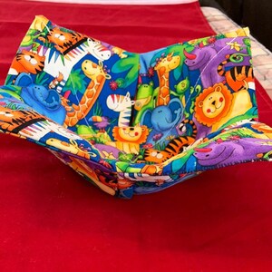 May include: A colorful fabric bowl with a jungle animal print. The bowl is made of quilted fabric and has a square shape. The fabric features a variety of animals, including giraffes, elephants, tigers, and zebras.