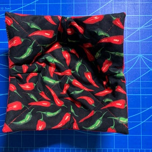 May include: A square, quilted bowl cozy with a black background and a pattern of red and green chili peppers. The cozy has a red border and is designed to hold a bowl of hot food.