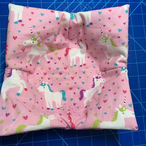 May include: A pink fabric pot holder with a unicorn and heart pattern. The pot holder is folded into a square shape.
