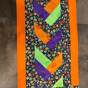 May include: A rectangular table runner featuring a central chevron pattern in alternating orange, purple, and lime green fabric strips. The border and background fabric display a black print with orange pumpkins, green leaves, and small white stars.