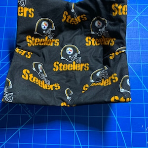 May include: Black fabric pouch with a repeating pattern of yellow Steelers logos and helmets. The pouch is folded and sitting on a blue cutting mat.