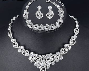 May include: A silver bridal jewelry set with a necklace, bracelet, and earrings. The set features a delicate, floral design with clear rhinestones.