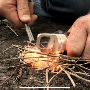 May include: A person using a ferro rod and striker to ignite a small pile of kindling. The kindling is made of small twigs and shredded material. The ferro rod is silver and rectangular. The scene is outdoors, with dirt and leaves in the background.