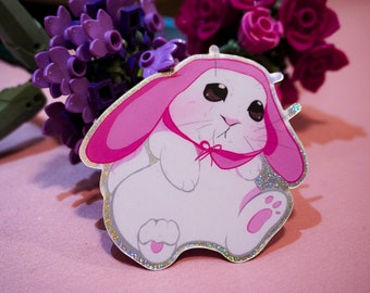 May include: A white bunny with pink ears and a pink bow, wearing a pink hooded sweater. The bunny is sitting on a pink surface with a glittery finish.