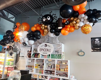 May include: A Halloween-themed balloon arch with black, orange, and white balloons, including a spider and pumpkin designs. The arch is hanging from the ceiling above a store with a sign that reads "Epicerie Grocery".