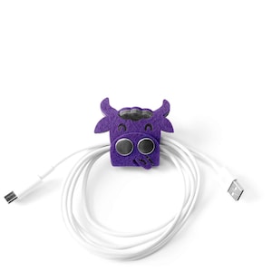 Könnte beinhalten: Lilafarbener Kabelorganizer aus Filz in Form einer Kuh mit schwarzen Augen und einem weißen Mund. Der Organizer hält ein weißes Kabel.