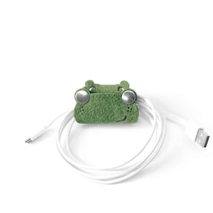 Può includere: Organizzatore di cavi in feltro verde a forma di rana con occhi a bottone argentati. L'organizzatore tiene un cavo bianco.