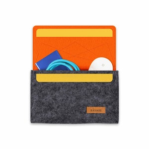 Könnte beinhalten: Eine graue Filzhülle mit offenem orangefarbenem Innenraum, die ein blaues Kabel, ein kleines blaues Notizbuch und einen weißen kreisförmigen Gegenstand enthält. Die Hülle hat einen gelben Akzent und ein braunes Lederetikett mit "RAVARE".