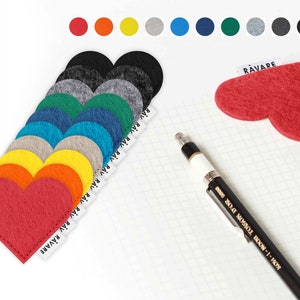 May include: A set of ten felt heart-shaped bookmarks in various colours, including red, orange, yellow, green, blue, and grey. The bookmarks are arranged in a row, with the red heart bookmark at the bottom. The bookmarks are labelled with the word "RAVARE".