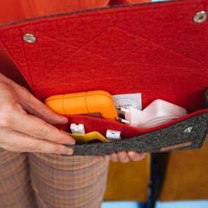 Könnte beinhalten: Eine graue Filztasche mit roter Filzfütterung. Die Tasche ist offen und enthält verschiedene Gegenstände, darunter ein kleines orangefarbenes Gerät, ein weißes Netzteil und ein gelbes Kabel.
