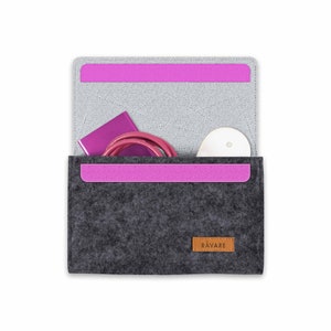 Könnte beinhalten: Eine graue Filztasche mit einem rosa Reißverschluss und einem braunen Lederanhänger mit der Aufschrift "RAVARE". Die Tasche ist geöffnet und enthält ein lilafarbenes Handy-Ladegerät, ein rosa Kabel und ein weißes Objekt.