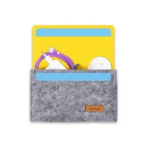 Könnte beinhalten: Eine graue Filztasche mit einer gelben Filzklappe, einem blauen Filmbesatz und einem braunen Lederanhänger mit der Aufschrift "RAVARE". Die Tasche ist geöffnet und enthält eine weiße kabellose Maus, ein violettes Ladekabel und ein weißes Ladegerät.