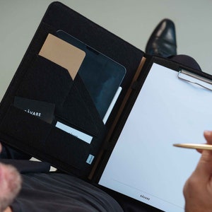 May include: A black felt portfolio with a built-in clipboard, a pocket for a tablet, and a smaller pocket for a notepad. The portfolio has the word "KAVARE" printed on the front.