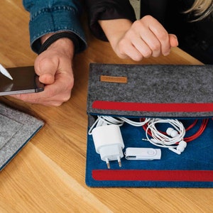 May include: A gray felt pouch with a red zipper and blue lining. The pouch is open and contains a white phone charger, a white USB drive, and white and red headphones.