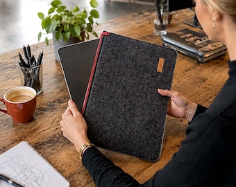 May include: A dark gray felt laptop sleeve featuring a vibrant red zipper closure along the side and a small rectangular brown leather-like brand tag attached to the upper right corner. The sleeve is rectangular with rounded corners and a textured finish.
