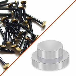 May include: Two round silver magnets, one stacked on top of the other. The magnets are on a white surface. A pile of black tacks with gold heads are in the background.