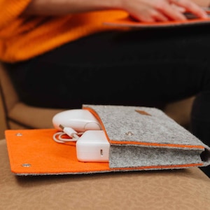 Könnte beinhalten: Eine graue Filztasche mit orangefarbenem Futter, die eine weiße kabellose Maus und ein weißes Netzteil enthält. Die Tasche ist geschlossen und liegt auf einer braunen Oberfläche.