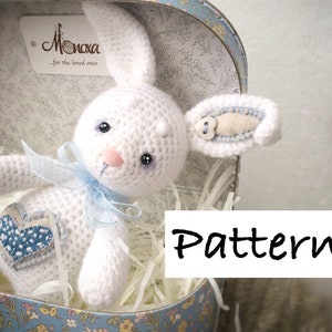 May include: White crocheted bunny rabbit toy with blue accents, a blue ribbon, and a heart-shaped patch. The bunny is sitting in a gift box with white shredded paper. The box has a blue and white floral pattern. The text "Pattern" is written in black on a white rectangle.
