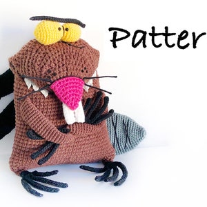 May include: Crocheted beaver plush toy with brown fur, black claws, a gray tail, and yellow eyes. The toy is sitting on a white surface with the word "Pattern" written in black above it.