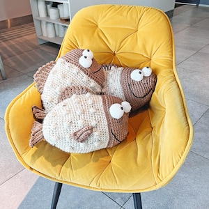 Op de afbeelding: Drie handgemaakte gehaakte vissenkussens in bruin- en crèmetinten liggen op een gele fluwelen stoel. De vissen hebben grote, witte wiebelogen en lachende monden. De stoel heeft een gecapitonneerde rugleuning en zwarte poten.