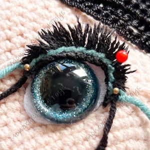 Puede incluir: Primer plano de un ojo de purpurina azul y verde con pestañas negras y una cuenta roja. El ojo está cosido en una tela de crochet rosa.