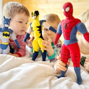 Puede incluir: Muñecos de superhéroes de Batman, Lobezno y Spider-Man hechos a ganchillo. El muñeco de Batman es azul y gris con un símbolo de murciélago amarillo. Lobezno es amarillo y azul. Spider-Man es rojo y azul.