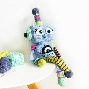 May include: A crocheted robot toy with blue, green, yellow, and purple accents. The robot has a friendly face with big eyes and a smile. It has a long, striped leg and a smaller, round leg.