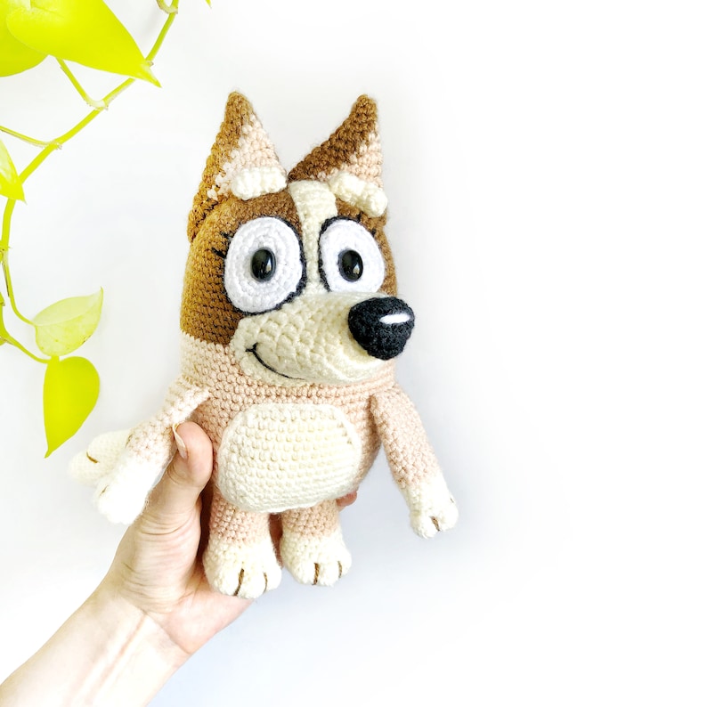 May include: A crocheted plush toy dog with brown and white fur, large eyes, and a black nose. The dog is smiling and has a friendly expression.